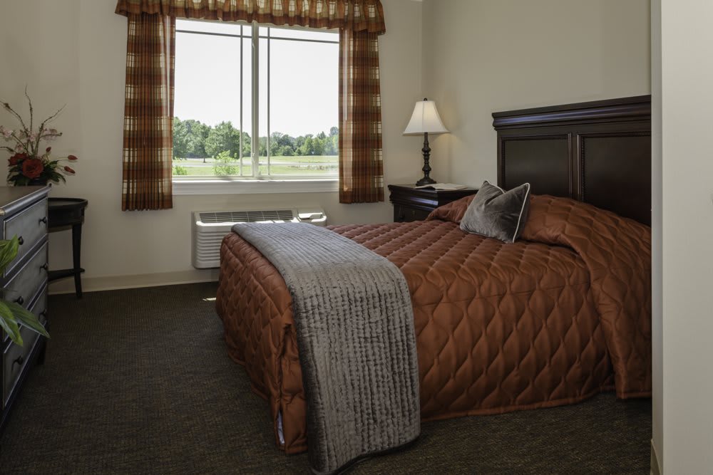 A cozy hotel bedroom with a brown quilted bedspread, a bedside lamp, and large window with plaid curtains.