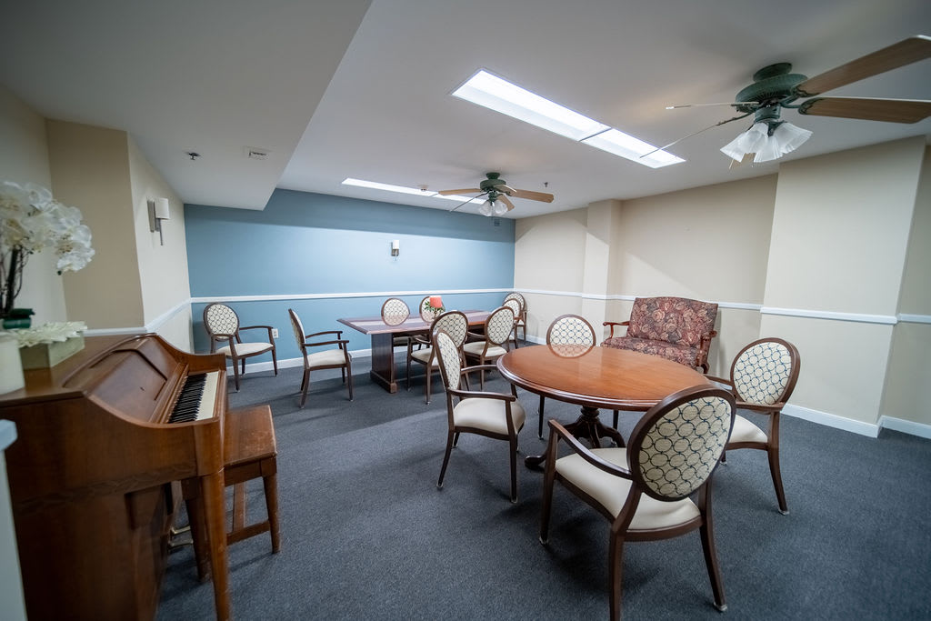 A cozy meeting room with multiple round tables and chairs, illuminated by natural light from the skylights, featuring a piano and ceiling fans.