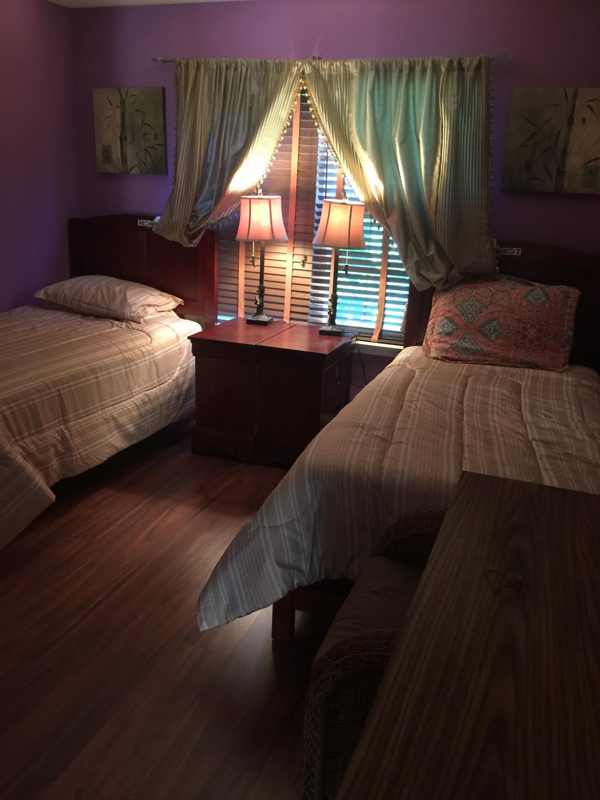 A cozy bedroom featuring twin beds, a nightstand with lamps, and soft lighting through the curtains.