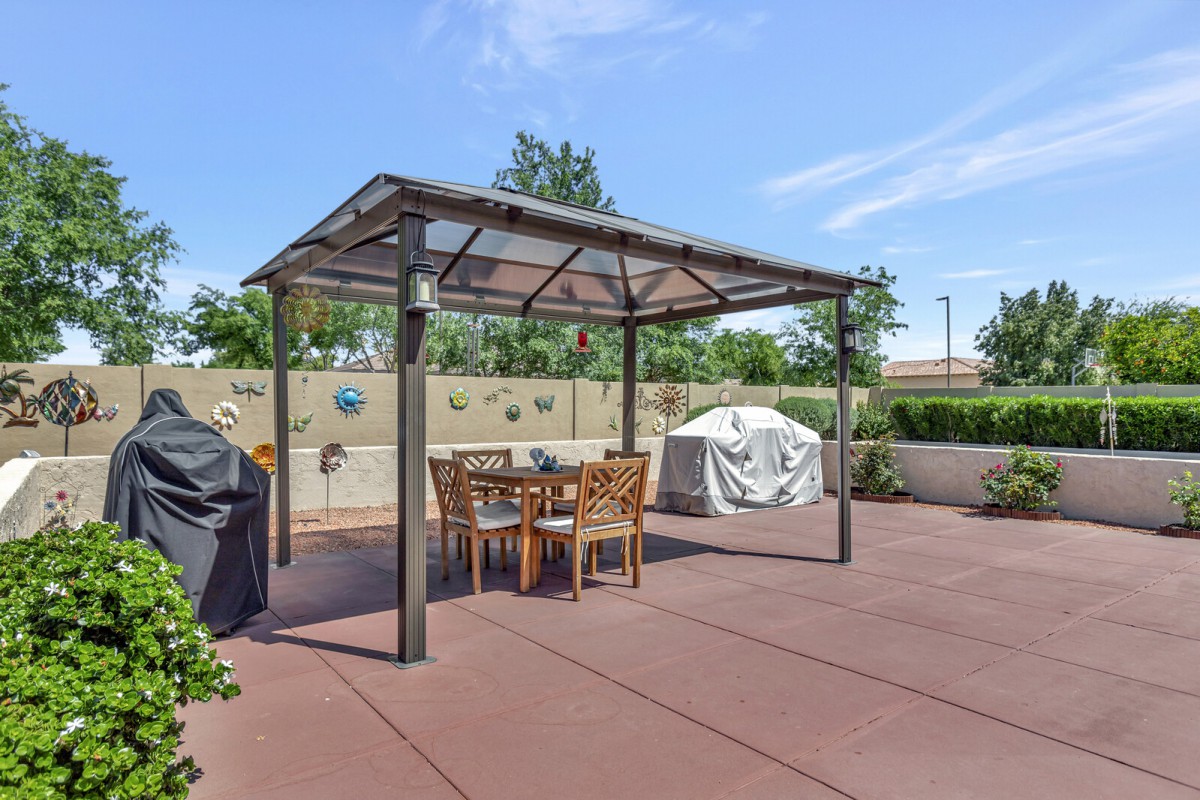 A backyard patio featuring a gazebo with outdoor dining furniture and a covered barbecue grill on a sunny day.
