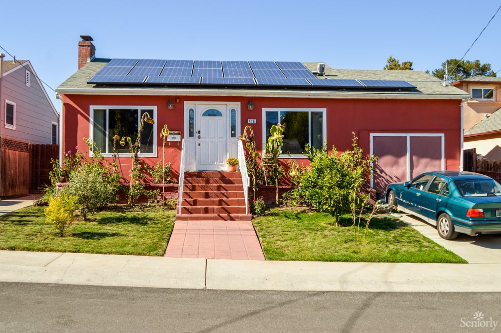 A red house with solar panels on the roof, featuring a front yard garden and steps leading to the entrance.
