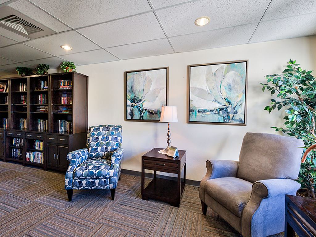 A cozy office interior featuring two chairs, a wooden bookshelf, artwork on the wall, and a small lamp on a side table.