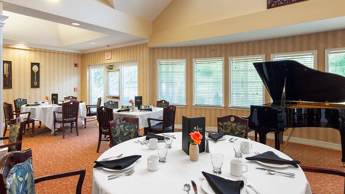 An elegant dining room with tables set for service, featuring white tablecloths, black napkins, and a grand piano by the window.