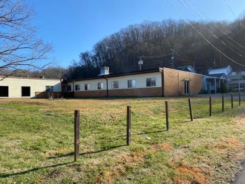 A single-story building with a brick facade is situated near a grassy field, bordered by a simple fence. Trees cover the hillside in the background under a clear blue sky.