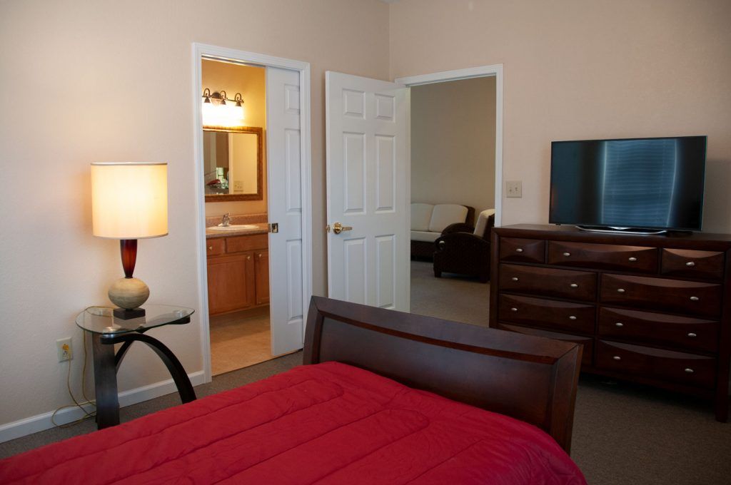 A cozy bedroom with a red bedspread, a lamp on a side table, and an open view to a bathroom and another room.