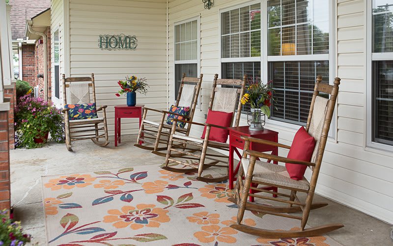 A cozy porch with wooden rocking chairs, red cushions, a floral rug, and vibrant flower arrangements.