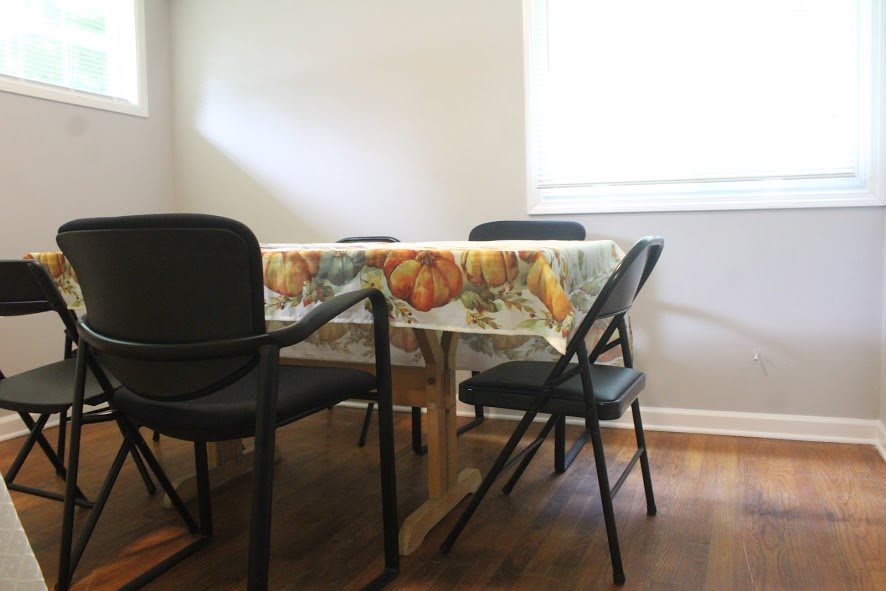 A dining area with a table covered by a patterned tablecloth and surrounded by chairs, illuminated by sunlight coming through the window.