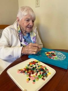 An elderly woman engaging in crafting with colorful buttons on a table.