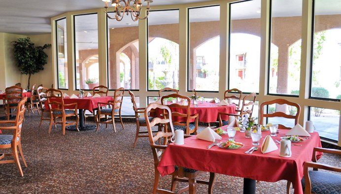 A cozy dining area with tables covered in red tablecloths, set against large windows.