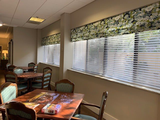 A cozy room with tables and chairs, featuring large windows with blinds.
