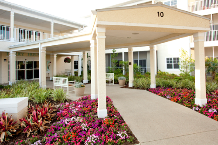 A well-maintained building entrance with colorful flower beds lining a concrete pathway, featuring white columns and a covered entryway.