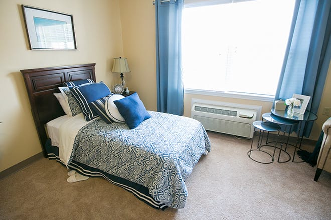A cozy bedroom with a single bed adorned in blue decor, featuring a lamp and a large window.
