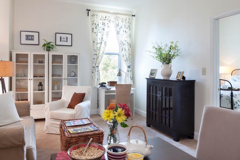 A cozy living room with a mix of modern and classic furniture, featuring a comfortable chair, a table with a flower arrangement, and a bookshelf.