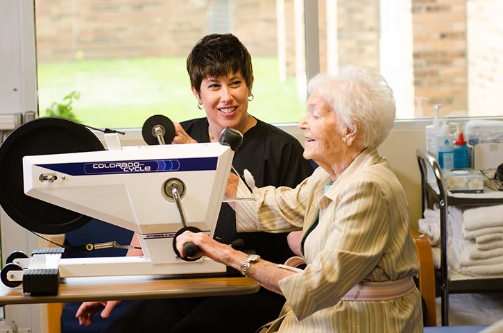 An elderly woman engages in physical therapy with assistance from a healthcare professional.