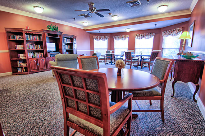 A cozy dining area with tables and chairs, featuring shelves and large windows with curtains.