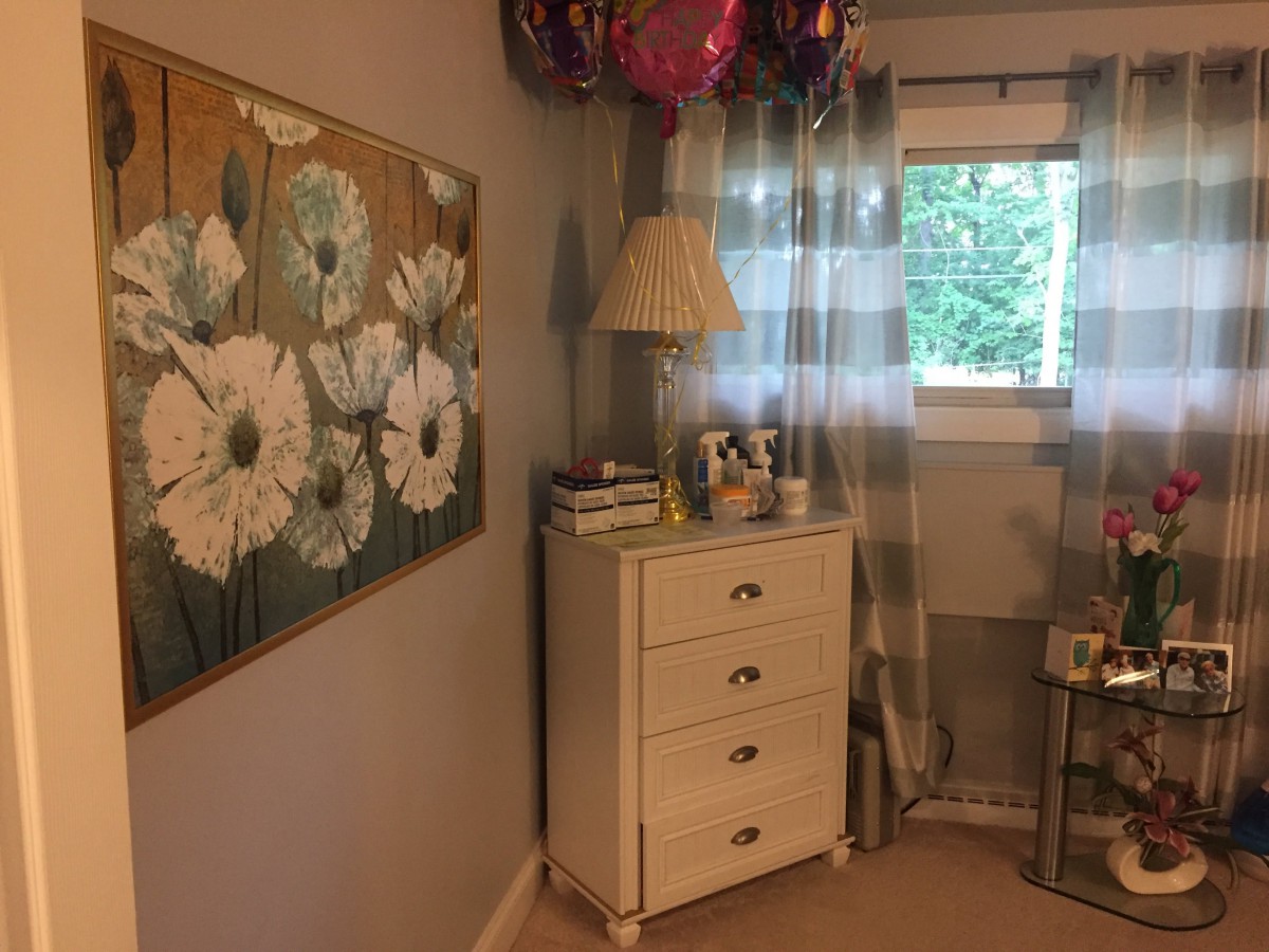 A cozy bedroom corner featuring a dresser with a lamp and balloons, alongside floral artwork and a window with curtains.