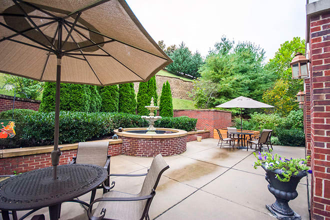 A tranquil outdoor patio area featuring tables and chairs under large umbrellas, with a decorative fountain surrounded by lush greenery.