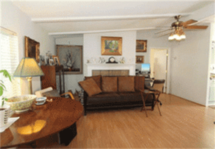A cozy living room with a brown sofa, wooden coffee table, and various artworks adorning the walls. A ceiling fan hangs above, and a computer desk is situated in the corner.