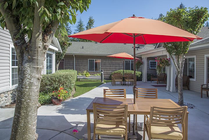 A sunny patio with wooden outdoor furniture and red umbrellas, surrounded by a well-maintained garden.
