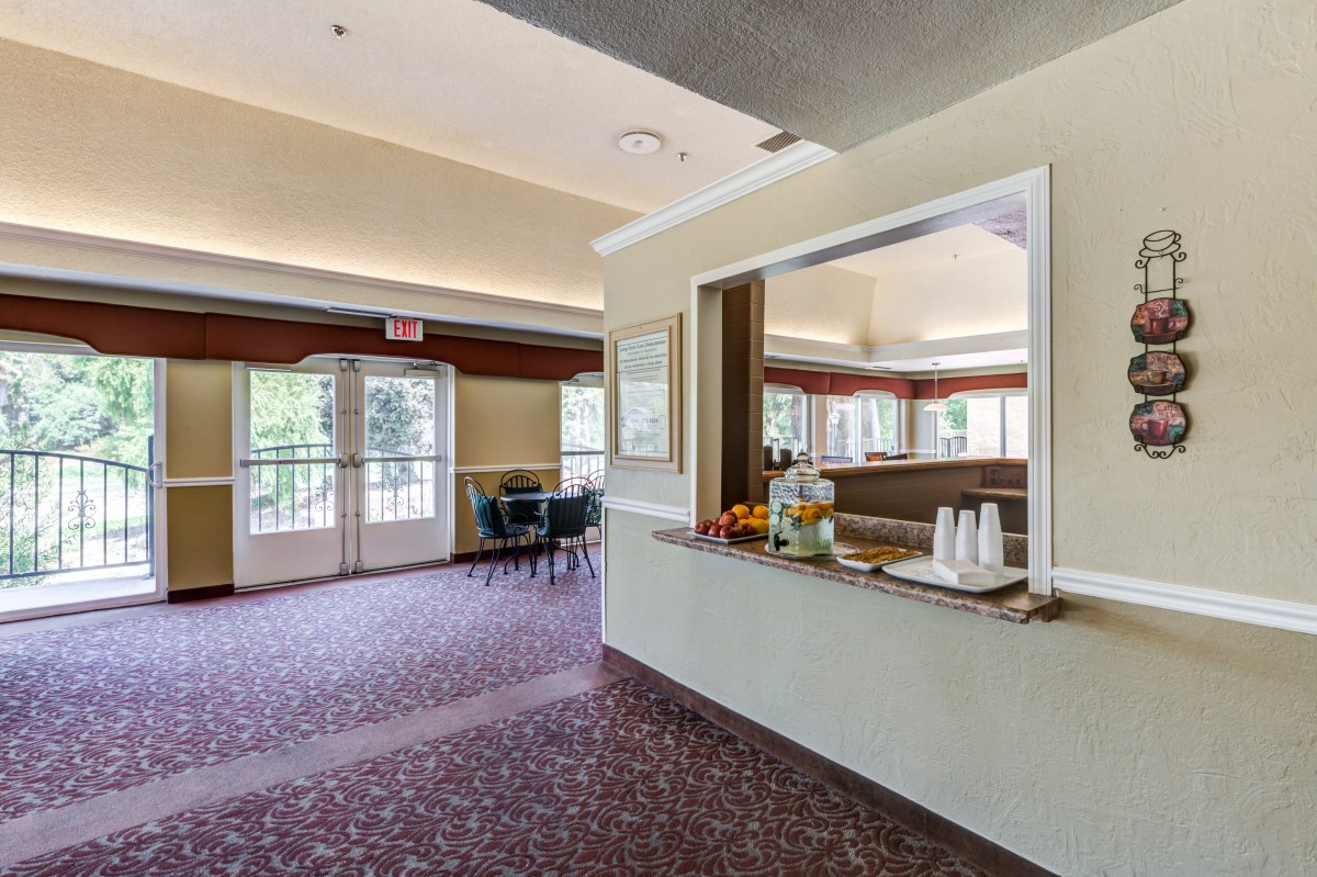 A spacious, carpeted room with a beverage station and seating area near large windows and double doors.