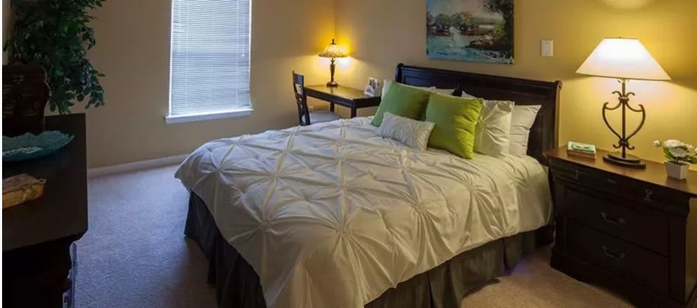 A cozy bedroom with a neatly made bed, accent pillows, and a well-lit desk lamp.