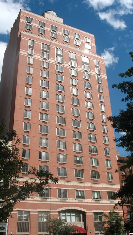 A tall brick building with numerous windows against a blue sky, framed by trees.