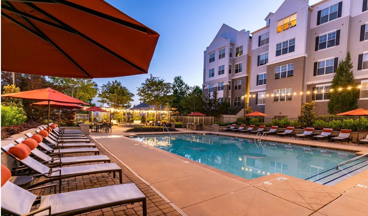 An outdoor swimming pool surrounded by sun loungers and umbrellas at an apartment complex during the evening.