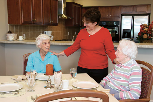 A caregiver engages warmly with two elderly women at a dining table in a senior living community.