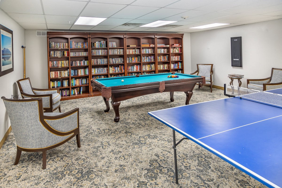 A cozy indoor recreation room featuring a billiard table, ping pong table, and a wall of bookshelves filled with books.