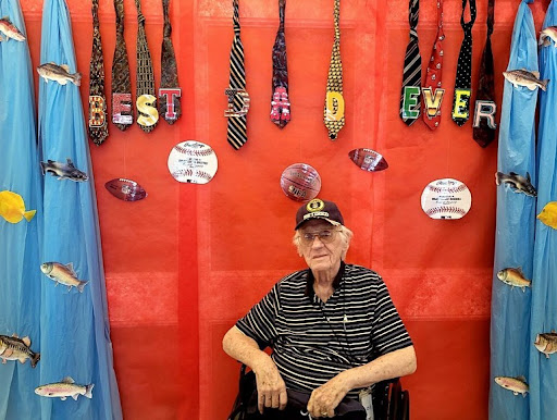 An elderly man sitting in a wheelchair with a display of ties behind him, celebrating 'Best Dad Ever'.