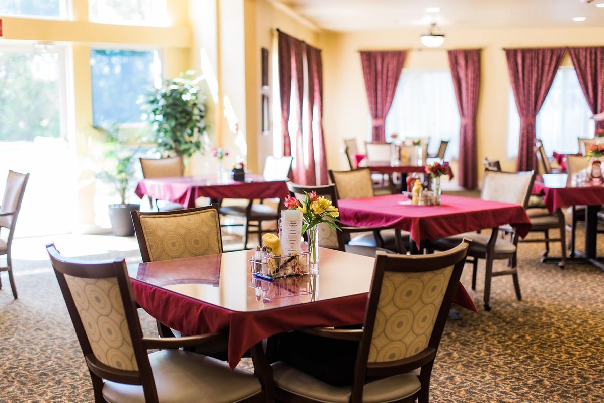 A peaceful indoor dining area with tables set with red tablecloths and chairs, decorated with curtains and plants.