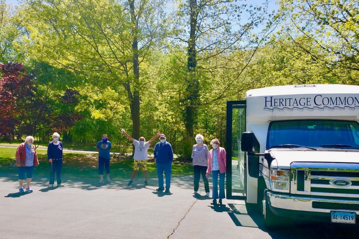A group of people standing near a bus in a sunny, tree-lined area.