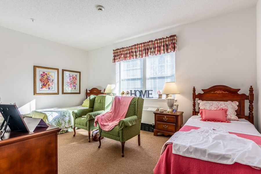 A cozy bedroom with twin beds, two green chairs, and a window with patterned curtains.