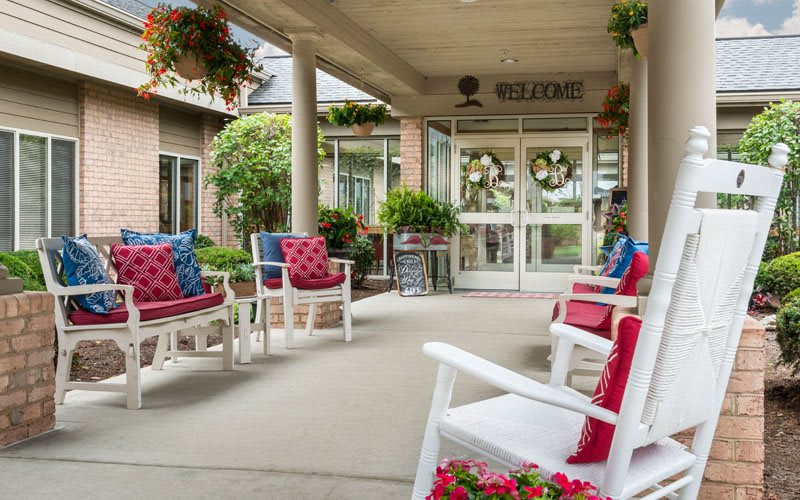 A welcoming porch area with rocking chairs adorned with decorative pillows, surrounded by plants at the entrance of a building.