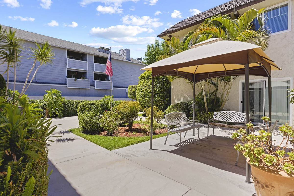 A peaceful courtyard featuring a gazebo with benches, surrounded by lush greenery and adjacent to an apartment building on a sunny day.