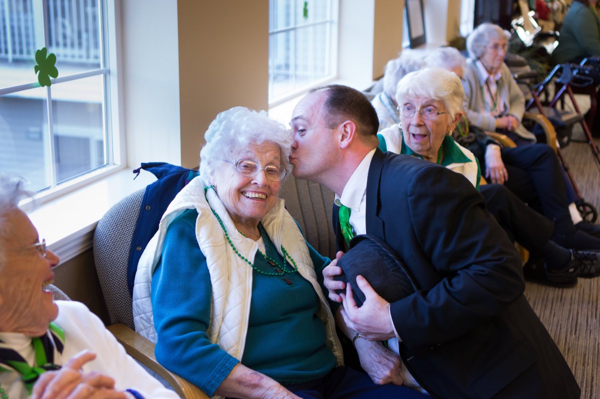 An elderly woman receives a warm kiss on the cheek from a man, surrounded by others indoors.