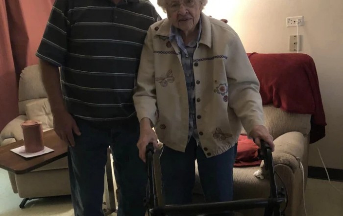 An elderly person using a walker indoors, accompanied by another individual.