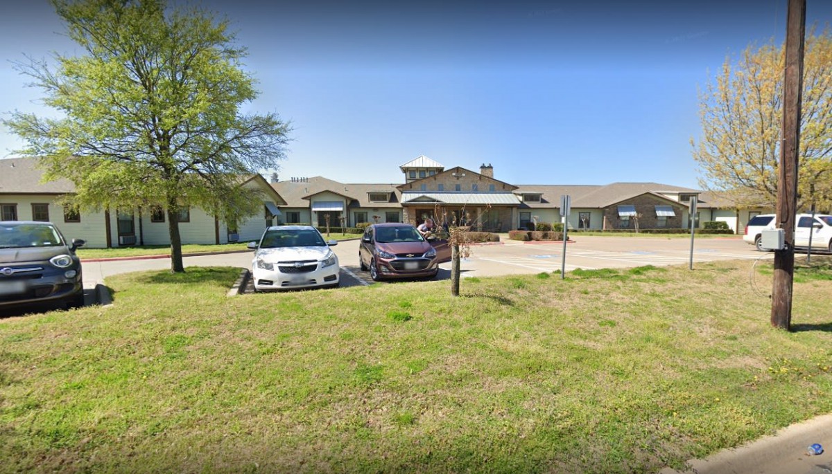 A building with a parking lot containing two cars, surrounded by grass and trees under a clear blue sky.