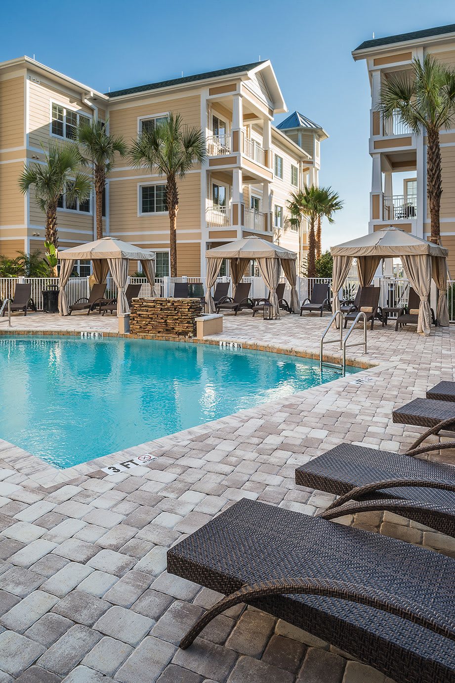 A luxurious resort area featuring a swimming pool surrounded by cabanas and lounge chairs, with palm trees in the background.