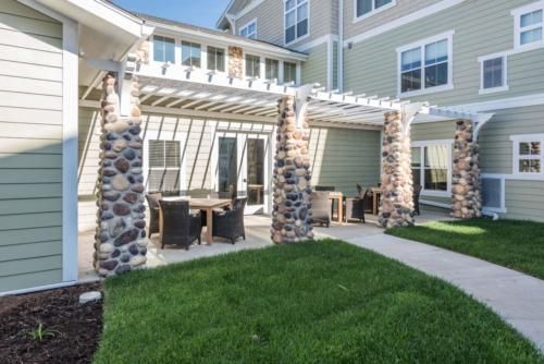 A modern patio with a pergola and outdoor furniture, complemented by stone columns and surrounded by a lush green lawn.
