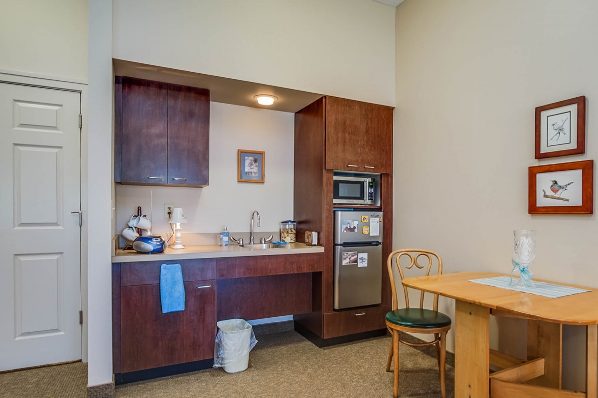 A cozy kitchenette area with wooden cabinets, a sink, and a microwave, adjacent to a small dining table and chair.