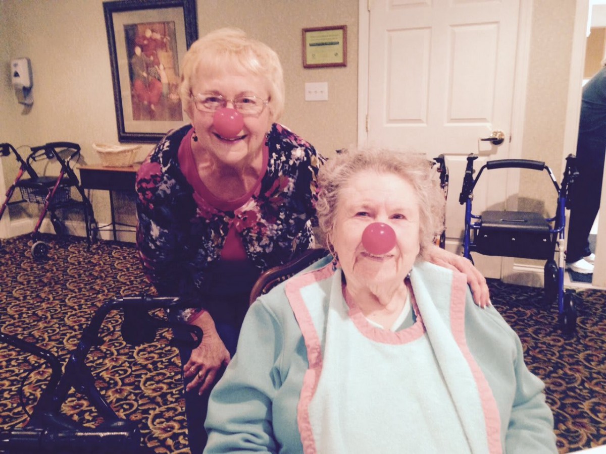 Two elderly women are smiling, with one playfully wearing a red clown nose, indoors.