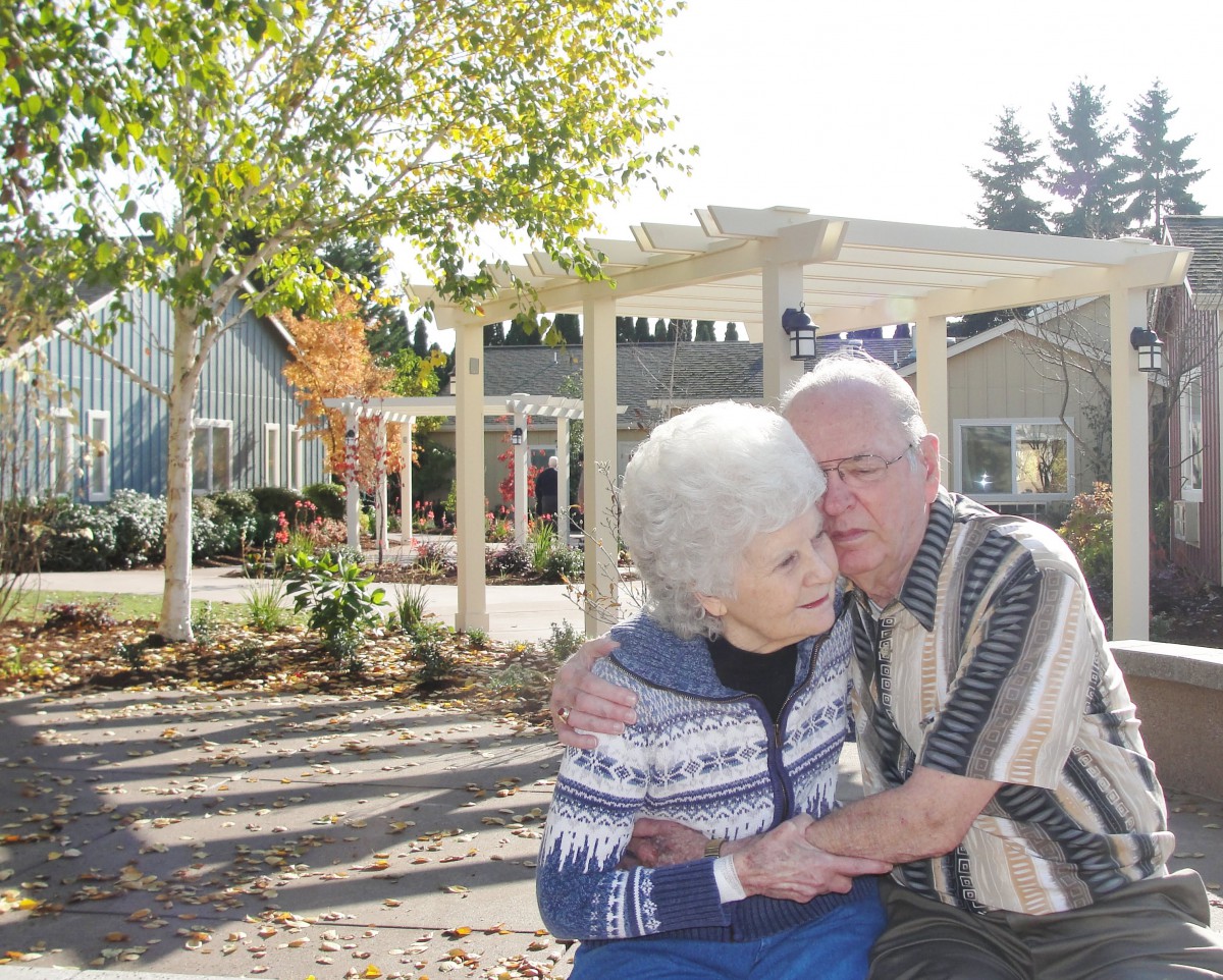 An elderly couple shares a tender moment in a garden setting.