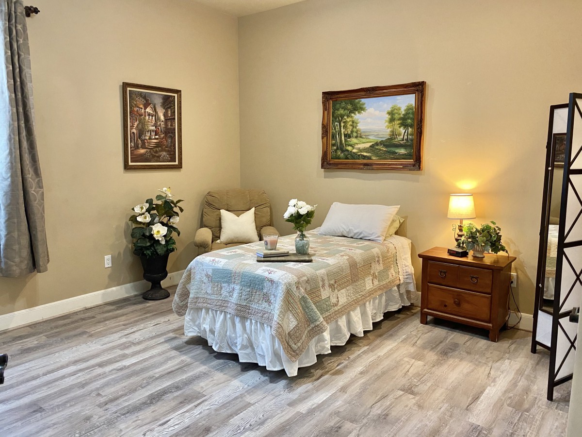 A cozy bedroom featuring a single bed, nightstand with lamp, decorative paintings, and floral arrangements.