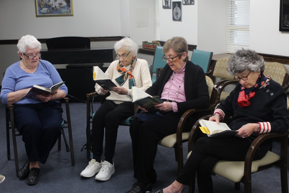 A group of elderly women engaged in a book club session, reading together in a cozy indoor setting.