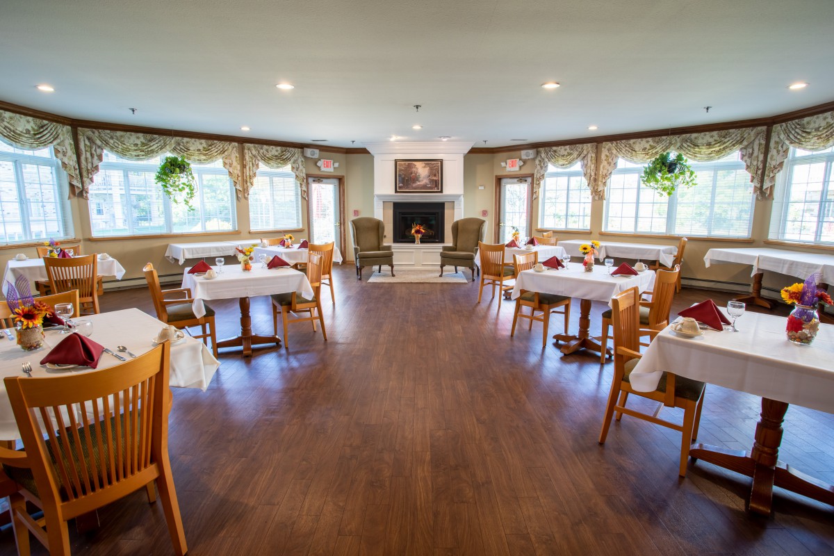 A cozy dining room with neatly arranged tables and chairs, featuring large windows and a central fireplace.