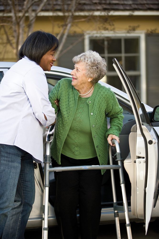 A caregiver assists an elderly woman with a walker as she gets out of a car.