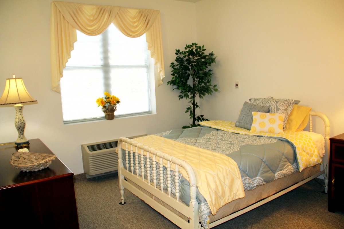 A neatly arranged bedroom featuring a bed with yellow and grey bedding, a window with decorative curtains, an indoor plant, and flowers on the windowsill.