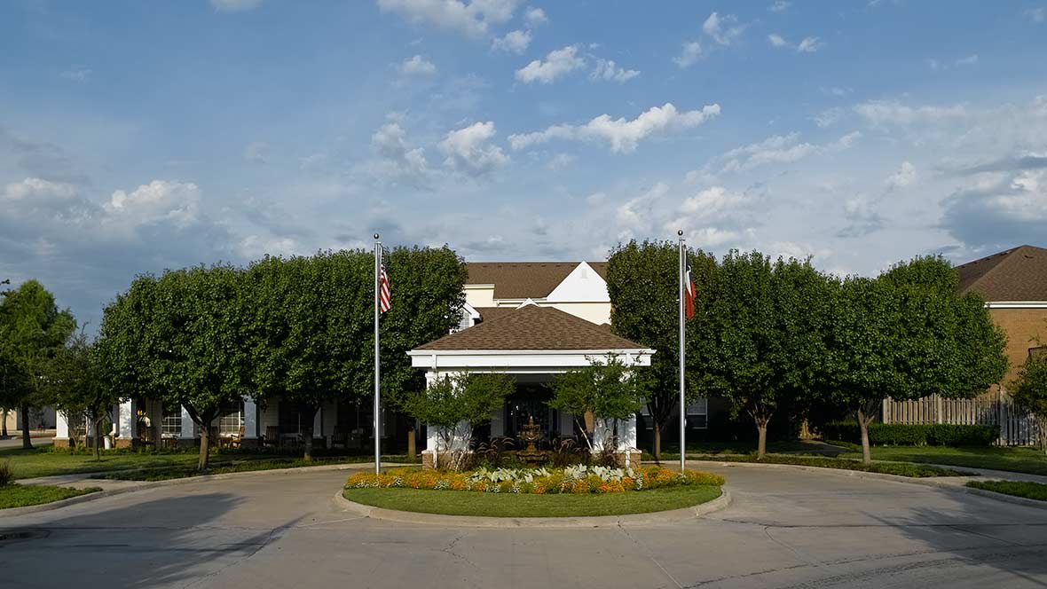 A building with flags in front, surrounded by trees and a landscaped roundabout under a partly cloudy sky.