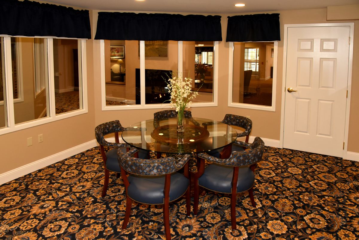 A cozy dining room with a round glass table and upholstered chairs, surrounded by large windows and decorated with a patterned carpet.